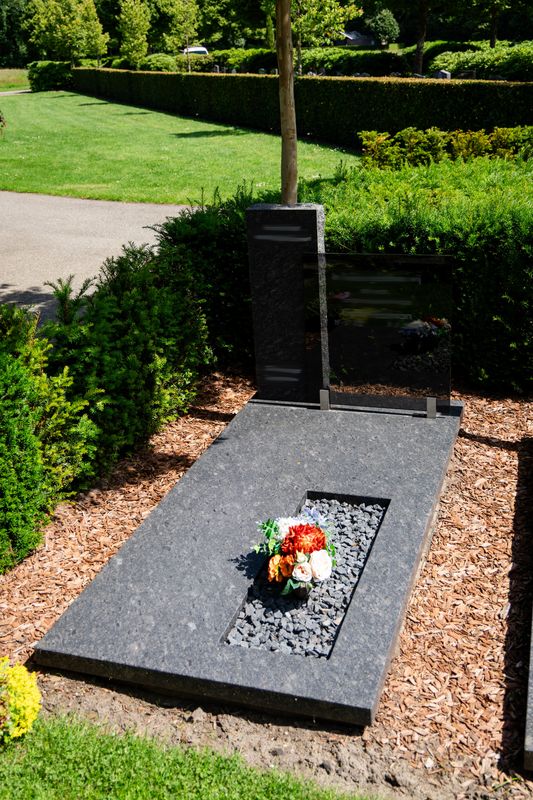 Eigentijds ligmonument van donker graniet met staande stele en grindvak met bloemen