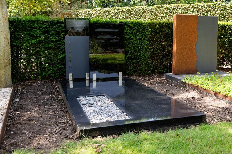 Strak grafmonument van donker graniet met grindvak, naast een monument van cortenstaal